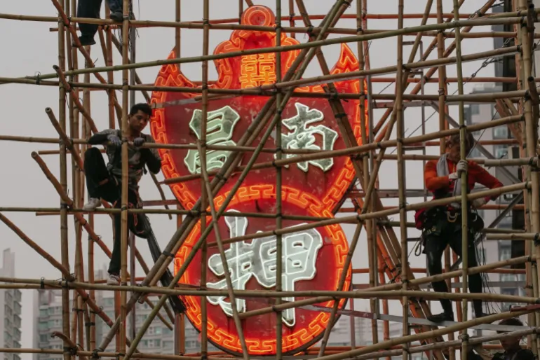 workers stand on bamboo next to a red neon sign