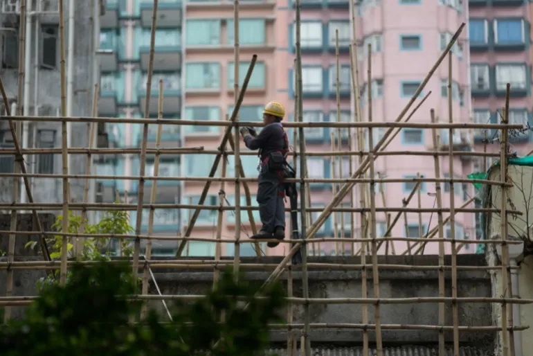 a worker in a hard hat stands on bamboo scaffolding near a high rise building