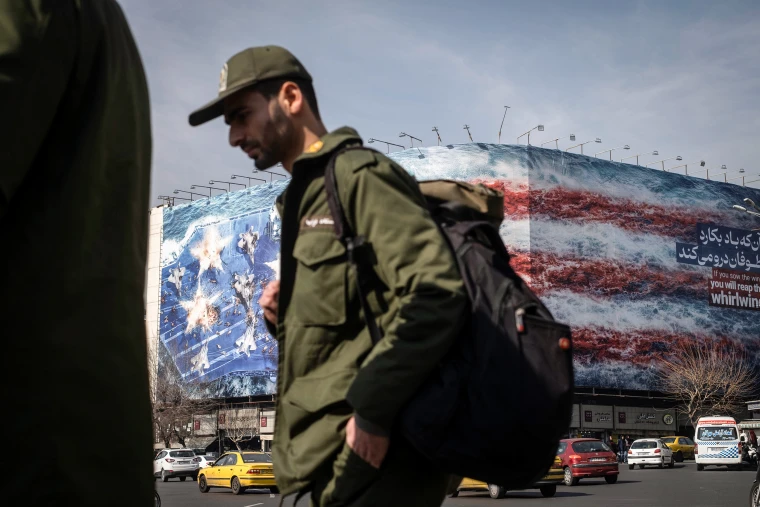 A soldier wearing green fatigues walks in front of a large building wrapped in a graphic bearing the stars and stripes of the American flag.