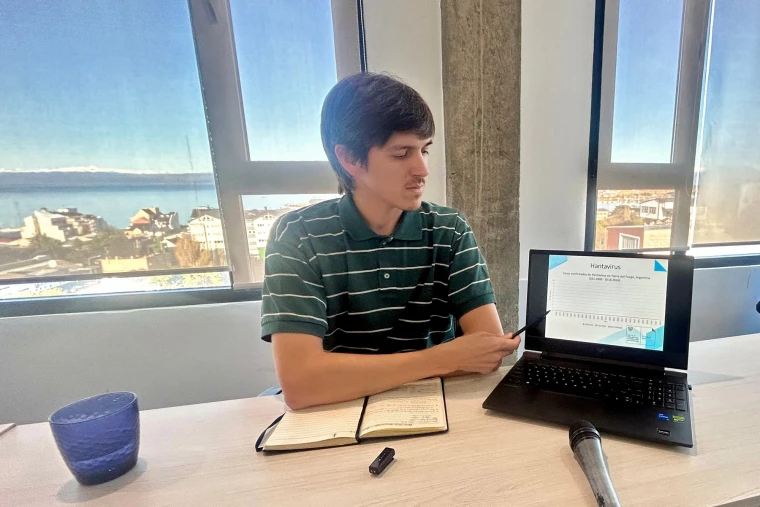 Juan Petrina sits at a table in front of windows and points to a laptop screen with a pen as his arms rest on a notebook