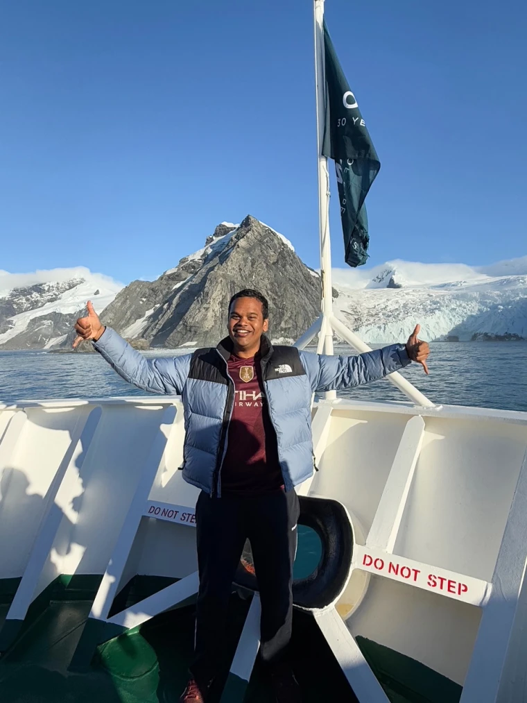Basanth Sadasivan poses with two thumbs up on the deck of a cruise ship, mountains can be seen behind him
