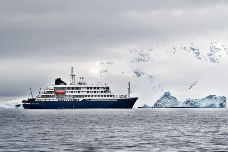 A cruiser ship in arctic waters with a large iceberg shrouded in clouds behind it